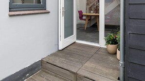 A narrow outdoor corridor with a wooden deck, a glass door leading to a room with a brick wall, and a small table with a chair. - Pakhuis aan de Zaan Wormerveer (Wormerveer)
