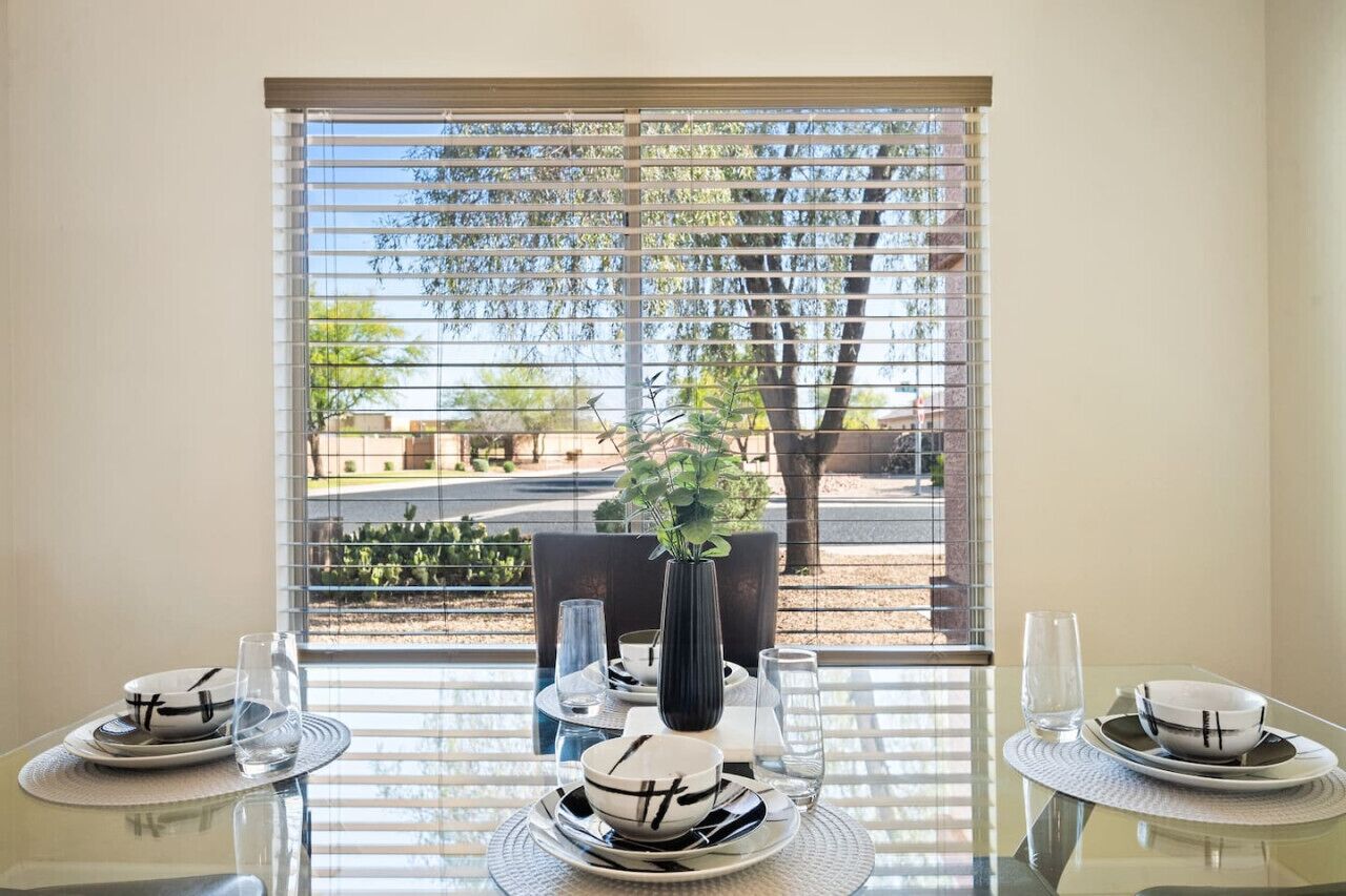Una mesa de comedor dispuesta con platos, tazones y vasos, ubicada cerca de una ventana con persianas.