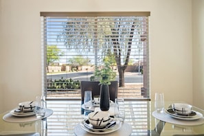 Une table à manger dressée avec des assiettes, des bols et des verres, placée près d’une fenêtre munie de stores.