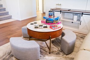 A living room with a round coffee table, two ottomans, and a bookshelf with board games.