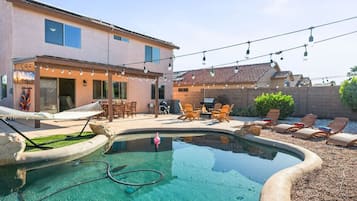 Un espace piscine doté de chaises longues et d’une petite piscine, le tout entouré d’un bâtiment avec une terrasse et des guirlandes lumineuses.