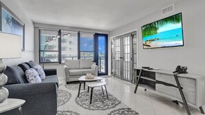 A modern living room with a grey sofa, a white coffee table, and a grey armchair. There is a large flat-screen TV mounted on the wall. - Cozy Beachfront 2 Bedroom Condo Beach Service 406 (Miami Beach)