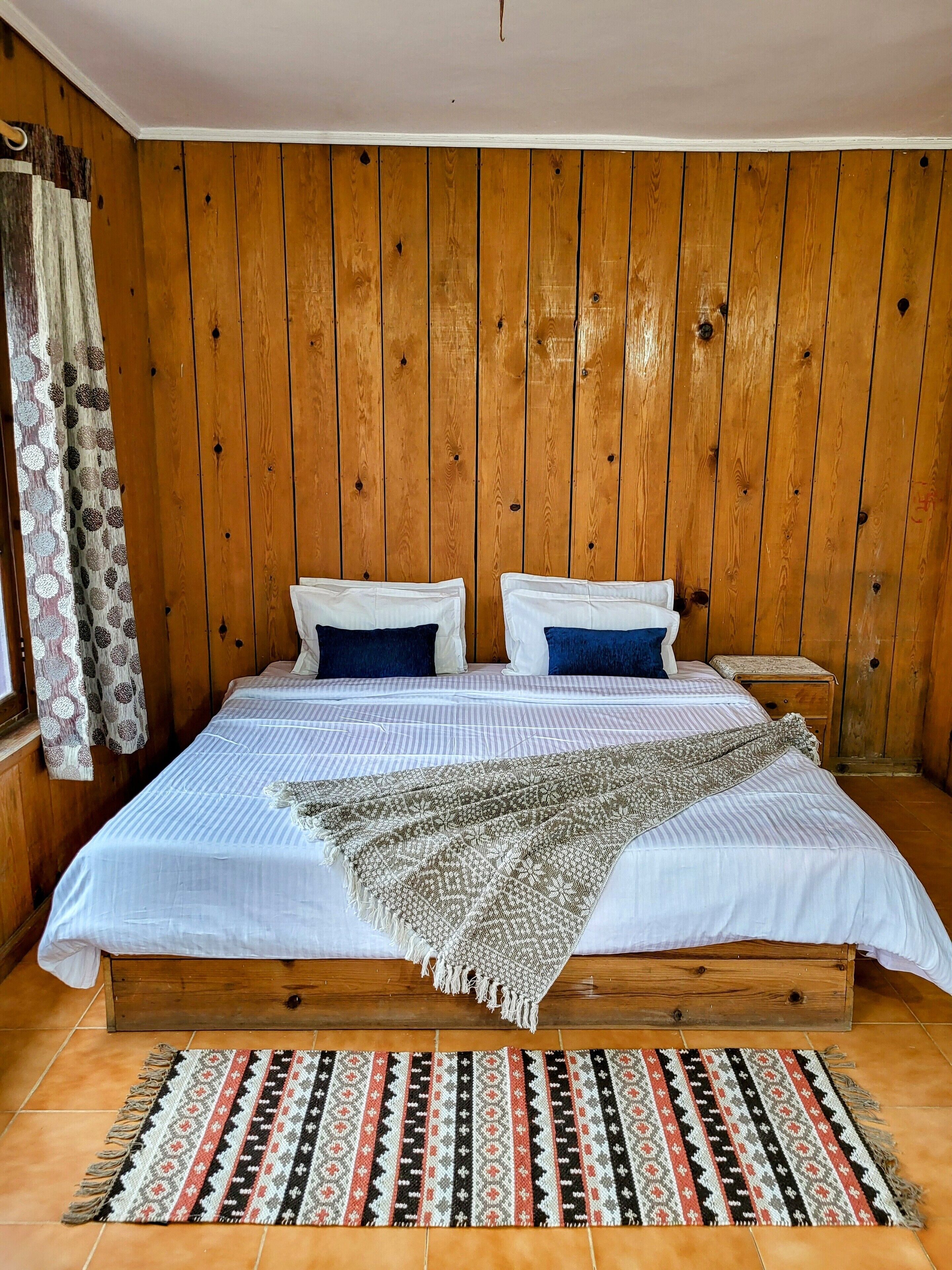 A wooden-paneled room with a bed, pillows, a nightstand, and a patterned rug.