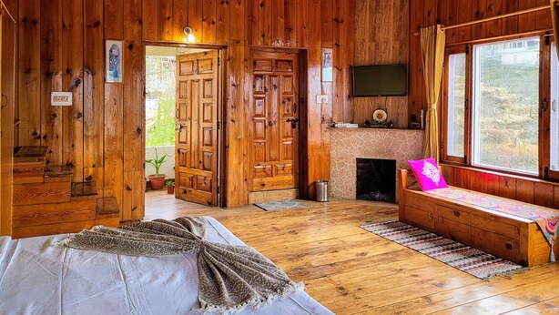 A wooden cabin interior with a bed, a fireplace, a TV mounted above it, and a window with a view of the outdoors. - Hartola Panorama Cottage (Kosiyakutoli)