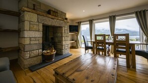 A cozy living room with a stone fireplace, wooden furniture, and a large window offering a view of the outdoors. - Avoca Lodge in the village of Onich (Fort William)