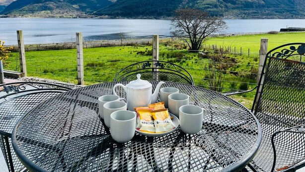 A table set with tea cups, a teapot, and snacks, overlooking a lake and mountains. - Avoca Lodge in the village of Onich (Fort William)
