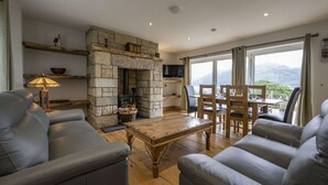 A cozy living room with a stone fireplace, leather sofas, a wooden coffee table, and a dining area with a view of the mountains. - Avoca Lodge in the village of Onich (Fort William)