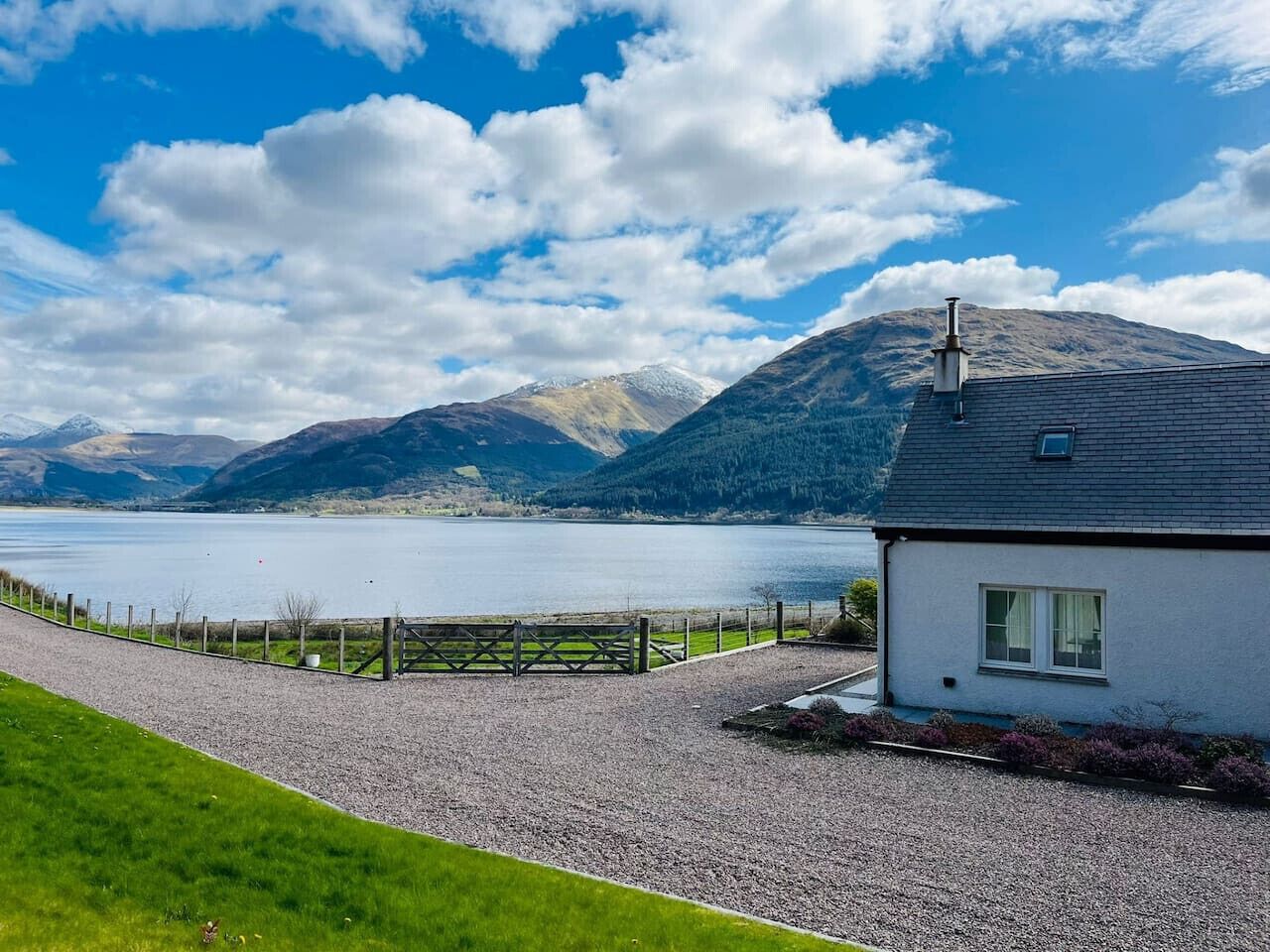 A house with a gravel driveway, a wooden fence, and a view of a lake and mountains.