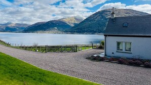 A house with a gravel driveway, a wooden fence, and a view of a lake and mountains. - Avoca Lodge in the village of Onich (Fort William)