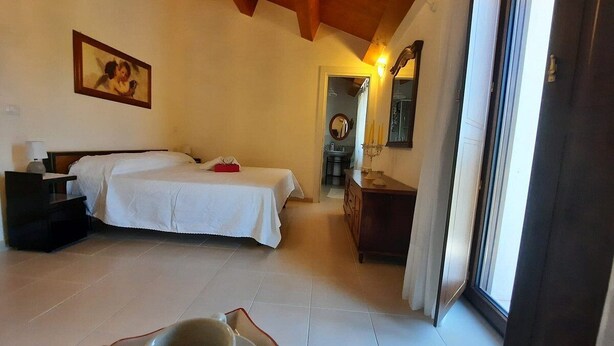 A bedroom with a bed, bedside table, and a small table with a cup. There is a door leading to another room and a window with a view. - B&B Santo Stefano (Campobasso)