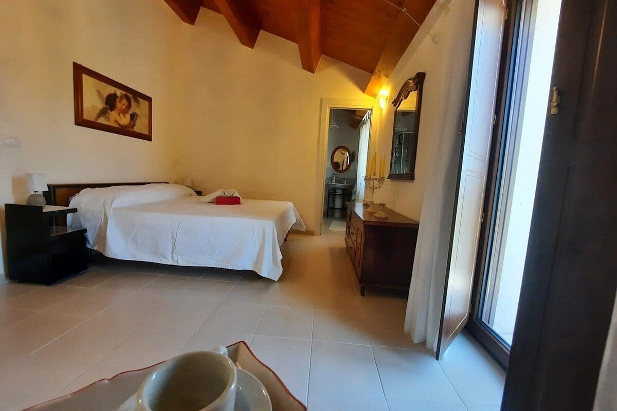 Un dormitorio con una cama, una mesita de noche y una pequeña mesa con una taza. Hay una puerta que da a otra habitación y una ventana con vistas.