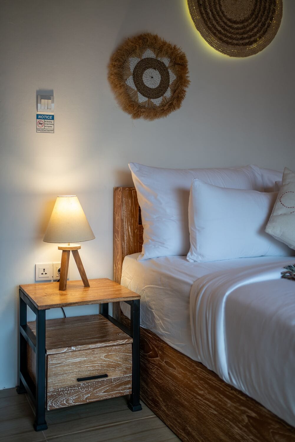 A neatly made bed with white linens, a wooden bedside table with a lamp, and a wall-mounted decorative item.