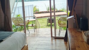 Een gezellig vakantiehuisje met een grote schuifpui die uitkomt op een terras met uitzicht op een meer en bergen. Binnenin is een bed en een houten tafel.