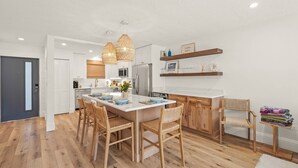 Une cuisine moderne avec des meubles blancs, un îlot central orné d’assiettes bleues, des chaises en bois et des fauteuils en osier.