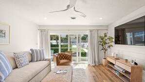 Un salon moderne comprenant un canapé, une table basse, un fauteuil, un ventilateur de plafond, une grande fenêtre avec des rideaux et un meuble TV en bois.