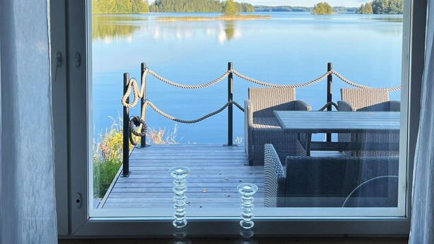 Une terrasse en bois avec une table et des chaises, donnant sur un lac paisible, agrémentée d’une barrière en corde et offrant une vue dégagée sur le paysage environnant.