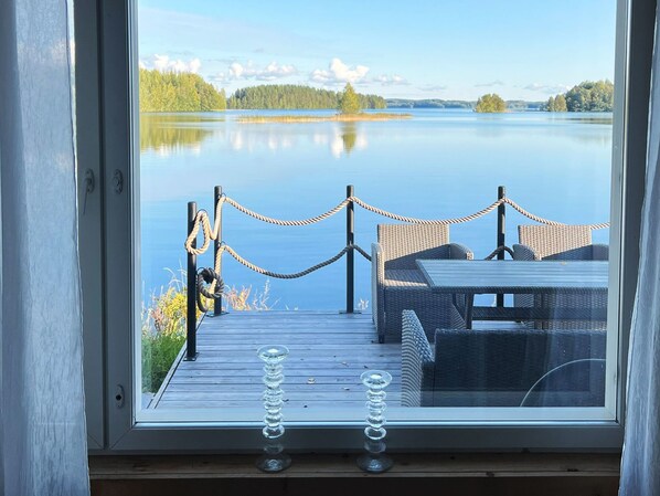 A wooden deck with a rope barrier, wicker furniture, and a clear view of a calm lake and distant trees.