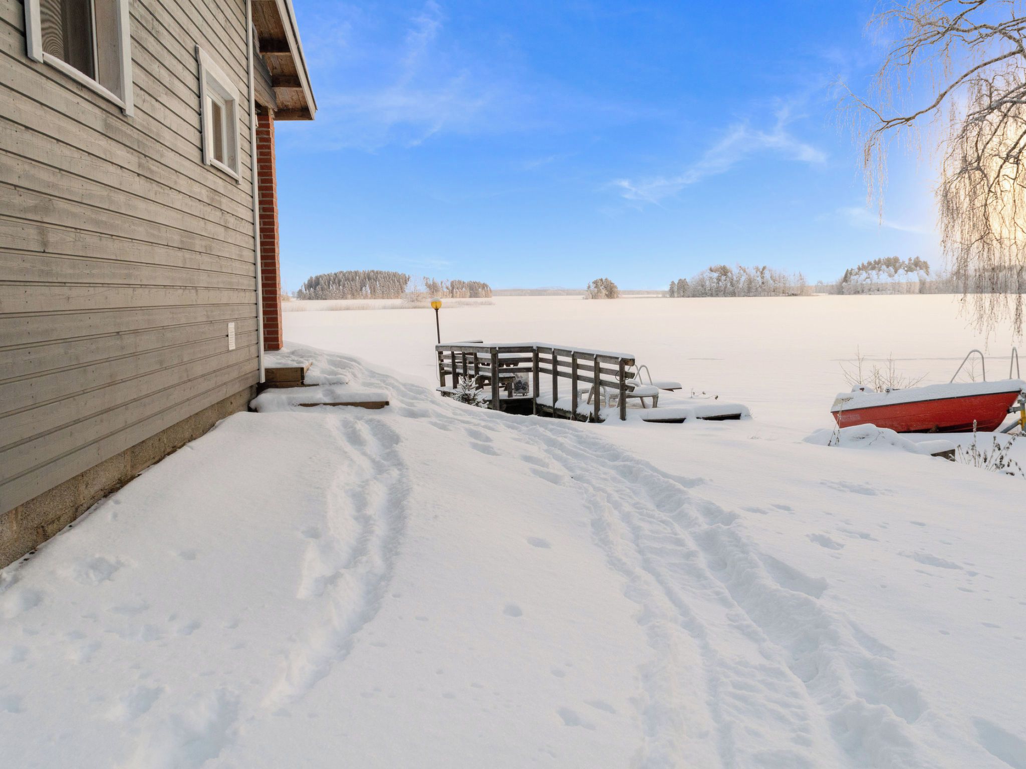 A snow-covered landscape with a wooden house, a snow-covered deck, and a red boat.