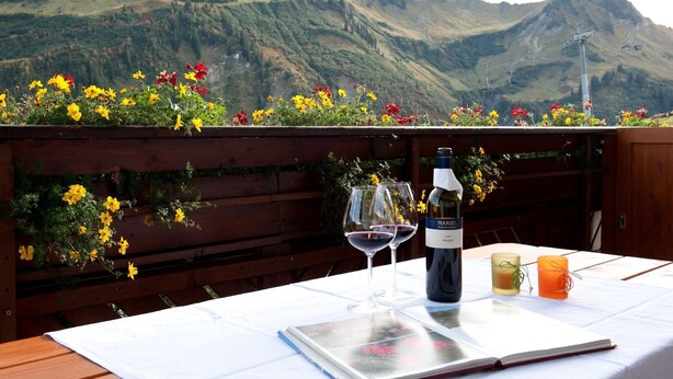 Tavolo apparecchiato all'aperto con una bottiglia di vino, due bicchieri e una candela su una tovaglia bianca.