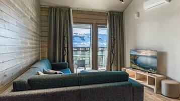 A modern living room with a sofa, a television, and a view of snow-covered mountains through large windows.