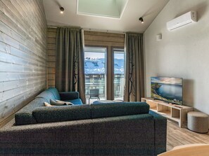En moderne stue med sofa, fjernsyn og udsigt til sneklædte bjerge gennem store vinduer.