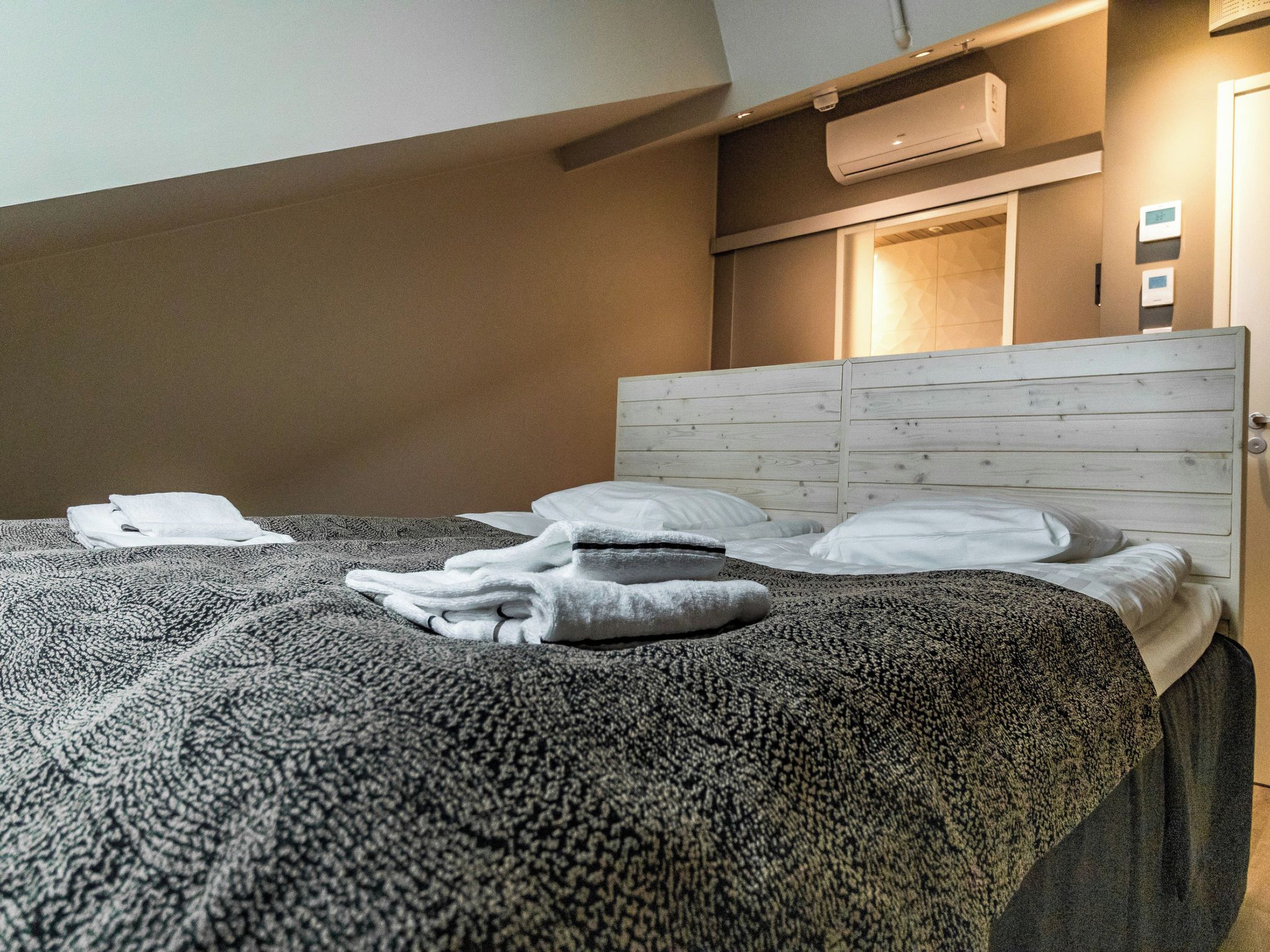 A hotel room with a bed, a headboard, a wall-mounted air conditioner, and a door with a digital display.