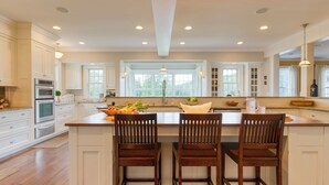 A spacious kitchen with white cabinetry, a large island with bar stools, and a view of the outdoors through large windows. - Private Estate, Guest House, Playroom, Gym, Theater | Sunny Pond by Avantstay (Austerlitz)