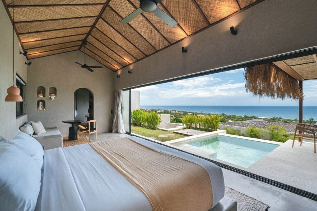 A bedroom with a large bed, a ceiling fan, a view of the ocean, and a small pool.