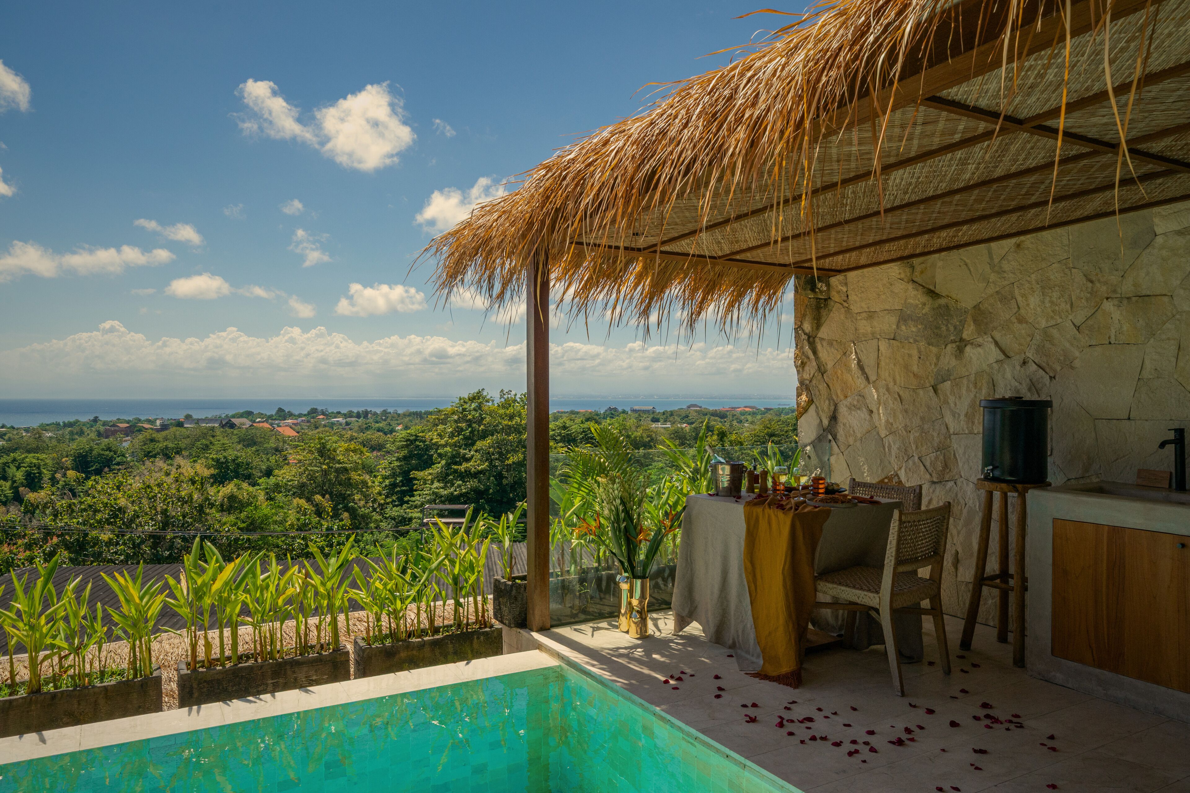 A pool with a view of the ocean, a thatched roof, and a stone wall.