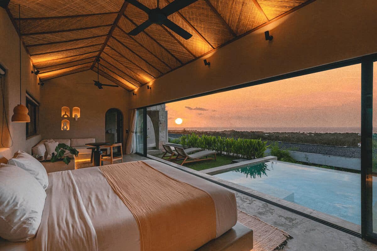 A bedroom with a large bed, a view of the sunset, a pool, and a dining area.