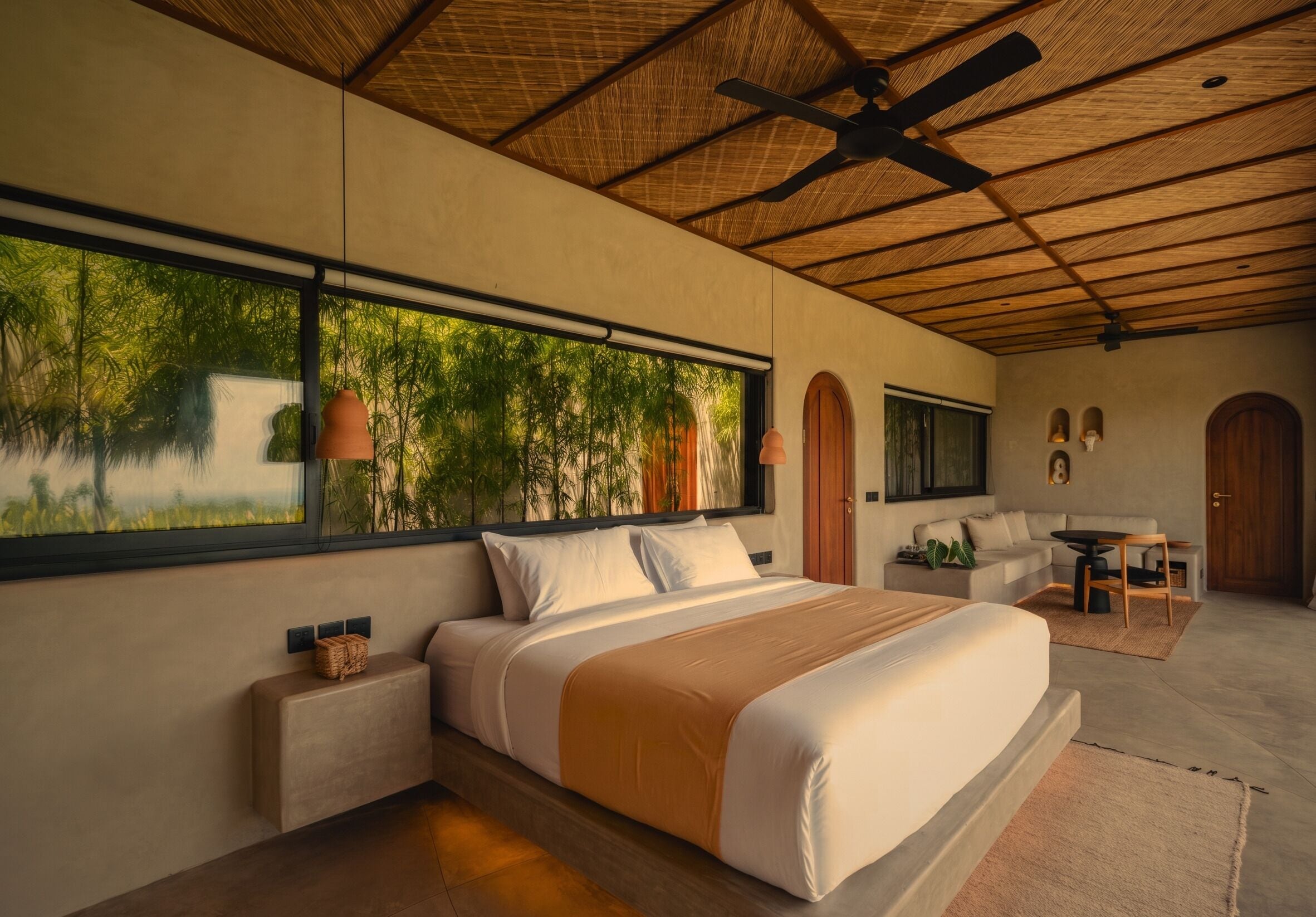 A modern bedroom with a large bed, a ceiling fan, a window with a view of greenery, and a small seating area with a table and chairs.