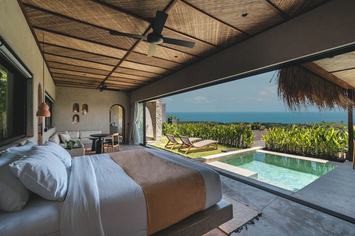 A bedroom with a large bed, a ceiling fan, and a view of a swimming pool and ocean.