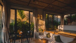 A modern living room with a sofa, dining table, and chairs, large windows with curtains, and a view of greenery outside. - SIVANA, Hotel Boutique with ocean or garden views - Adults only (Pecatu)