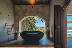 Un baño con paredes de piedra, un lavabo negro grande, una escalera de madera y vistas al mar a través de un arco.