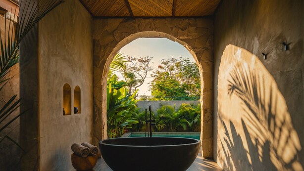 A spa room with a large black bathtub, a small wooden stool with towels, and a view of lush greenery through an arched doorway. - SIVANA, Hotel Boutique with ocean or garden views - Adults only (Pecatu)