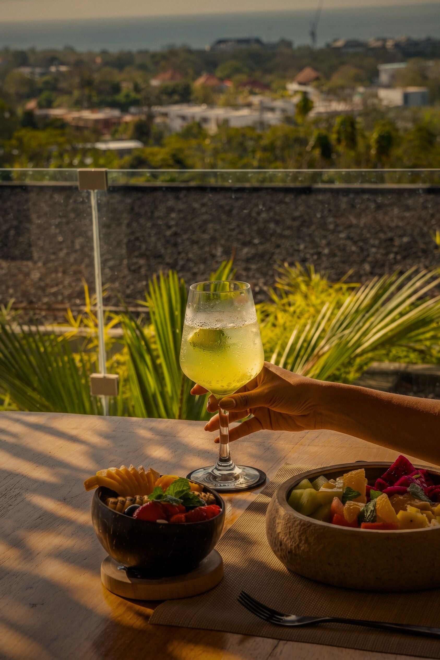A table with a fruit bowl and a glass of drink, a fork, and a view of greenery and buildings in the background.