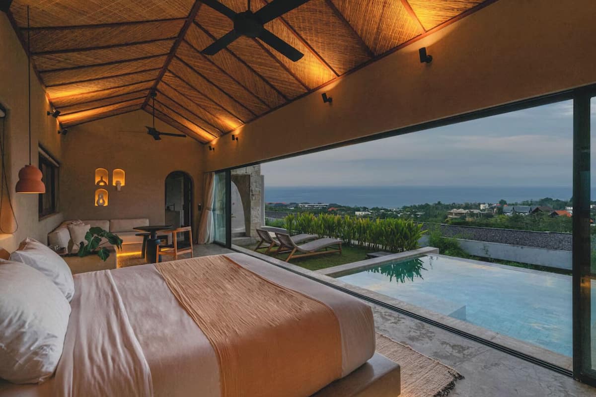 A bedroom with a large bed, a desk, and a view of a swimming pool and ocean.