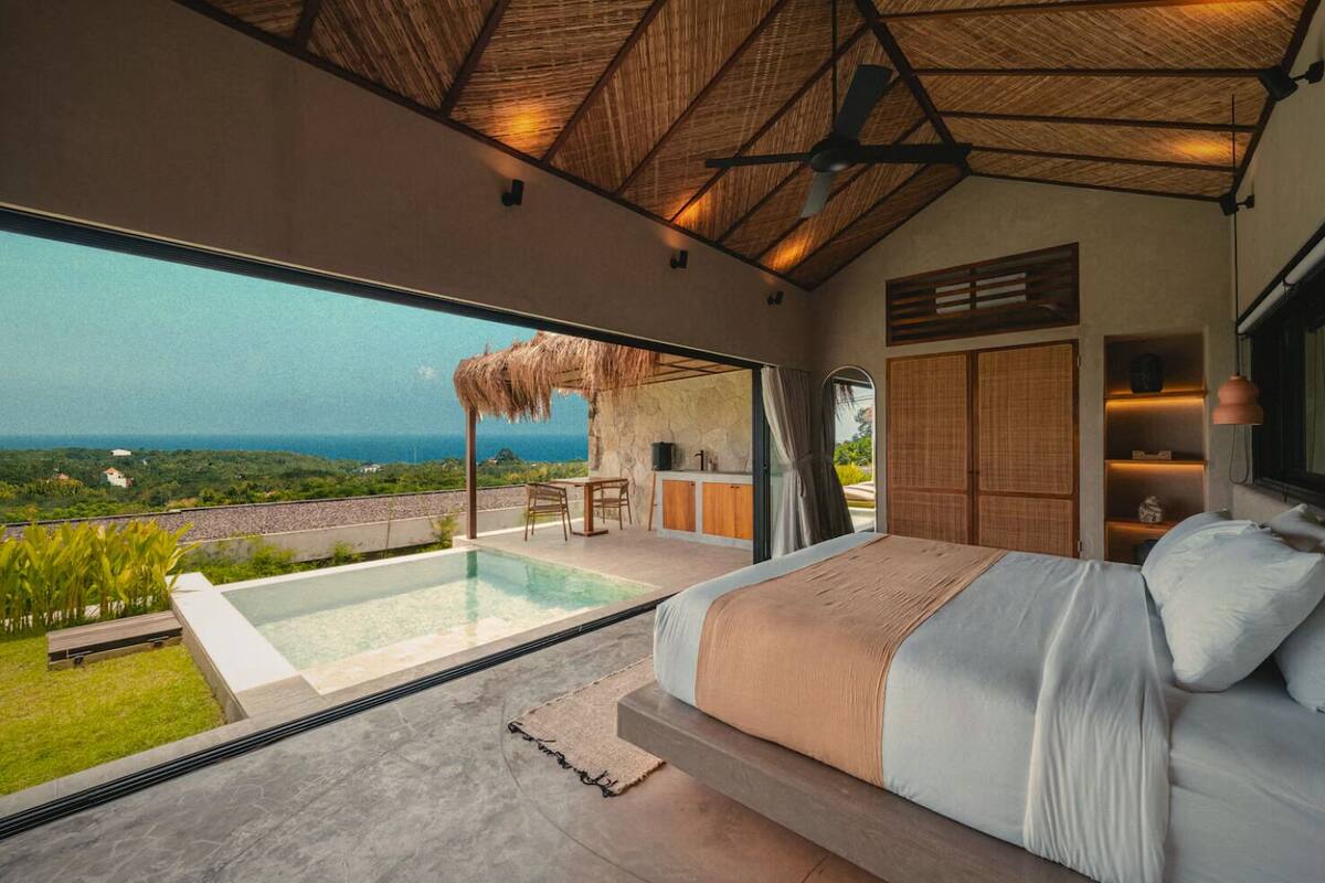A bedroom with a large bed, a view of the ocean, a small outdoor seating area, and a ceiling fan.