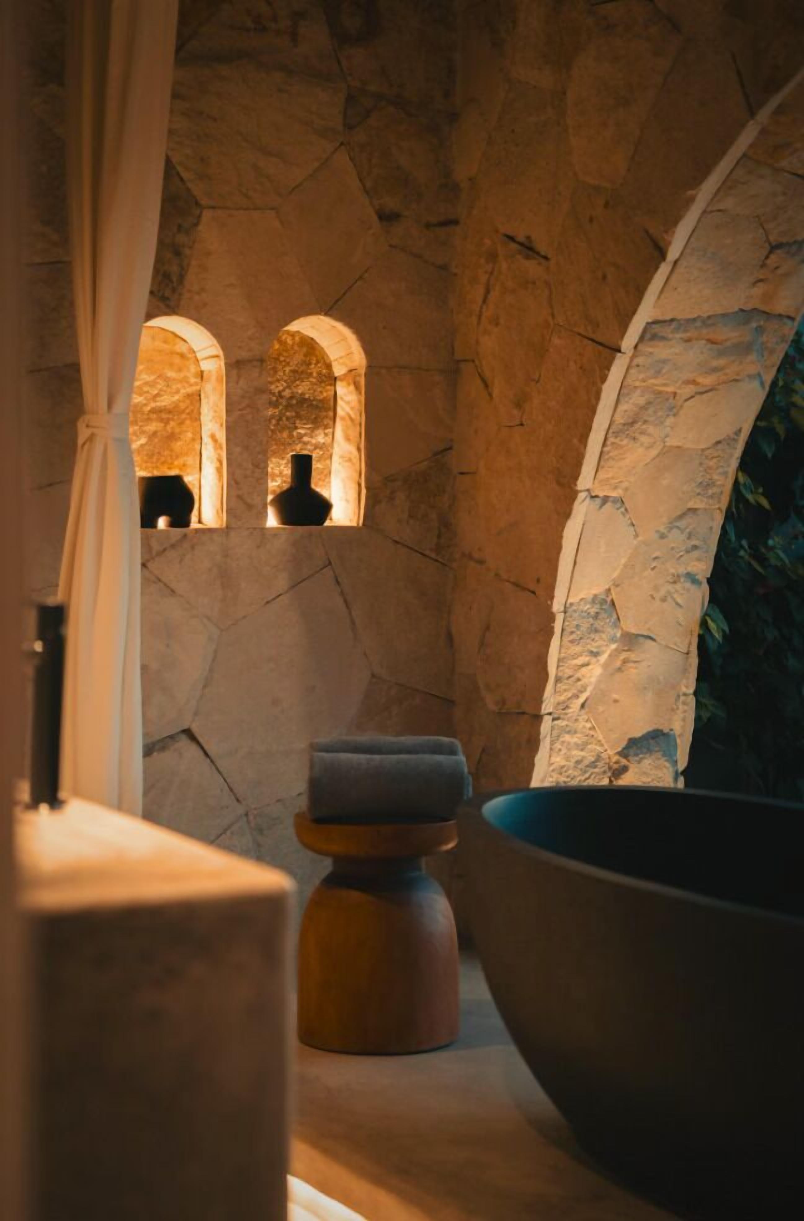 A stone wall with two arched windows, a large stone basin, and a wooden stool.