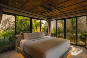 A large bed with a headboard, a ceiling fan, and a view of lush greenery outside.