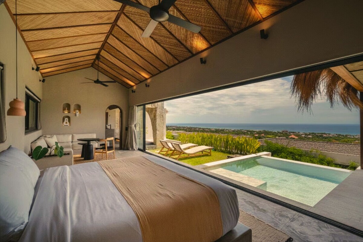 A bedroom with a large bed, a desk, a chair, a small table, a ceiling fan, and a view of the ocean.