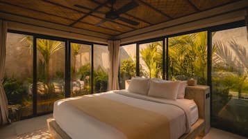 Une chambre moderne avec un grand lit, un ventilateur de plafond et de grandes fenêtres donnant sur un jardin tropical.