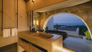 Un baño con una bañera grande, un lavabo y un espejo. Hay una pared de piedra y un arco que da a una vista.