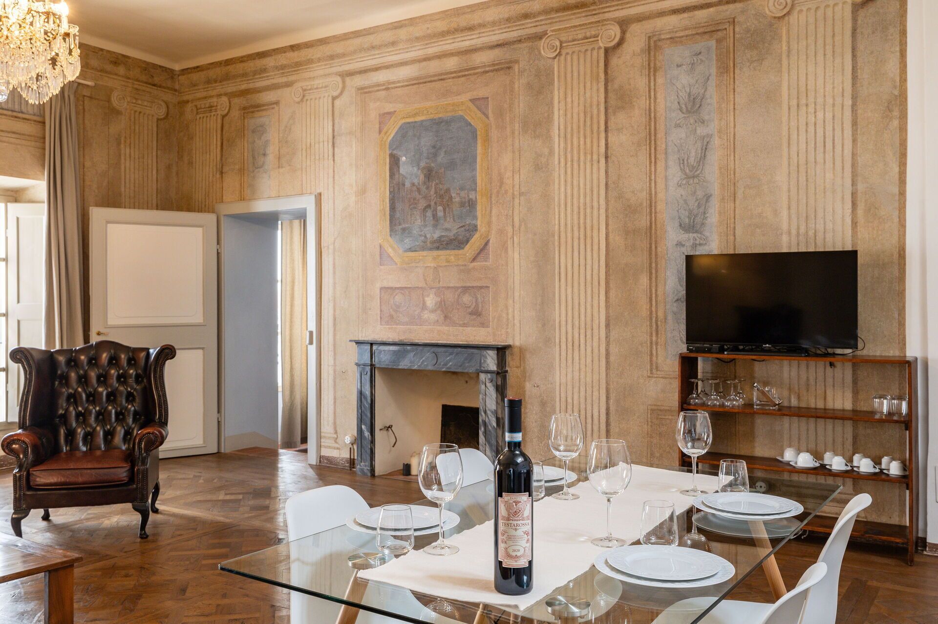 Un comedor con una mesa de vidrio, sillas blancas, una chimenea, un televisor sobre una estantería y un sillón clásico de cuero.