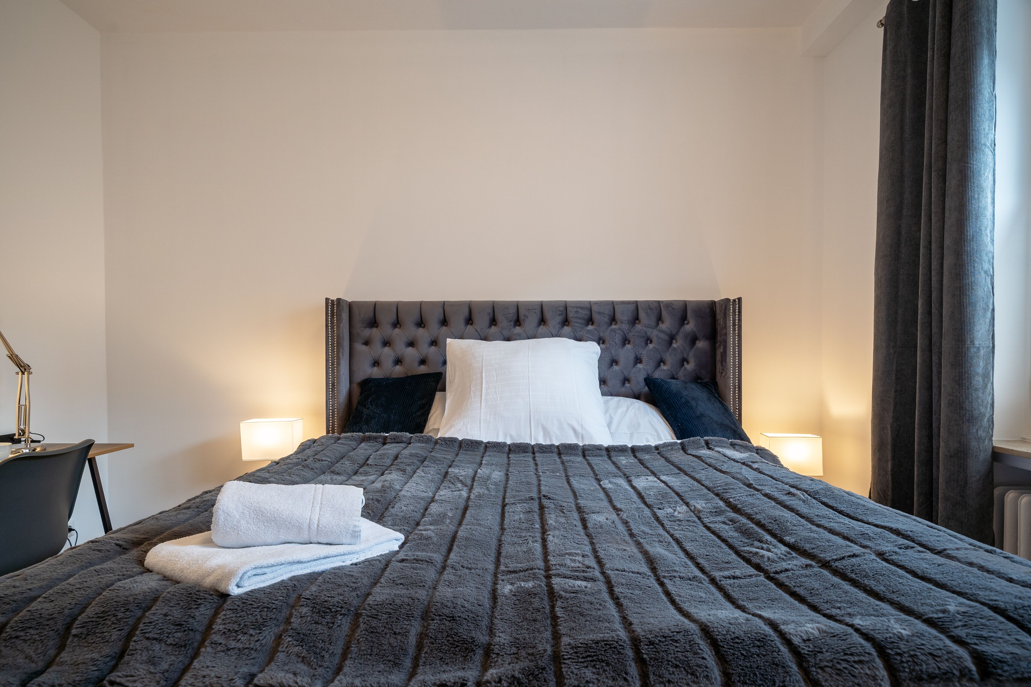 Una cama bien hecha con un cabecero capitonado, dos lámparas de noche y una toalla doblada sobre la cama.