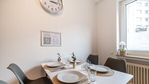 Comedor con una mesa blanca dispuesta para cuatro personas, con platos, cubiertos y copas. Un reloj está en la pared y debajo un certificado enmarcado.