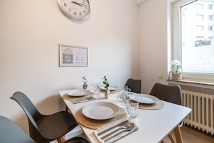 A dining area with a white table set for four, including plates, cutlery, and glasses. A clock is on the wall, and a framed certificate hangs below it.