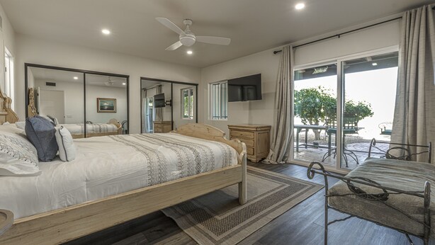 A bedroom with a bed, a ceiling fan, a TV, a window with curtains, and a balcony with outdoor furniture.