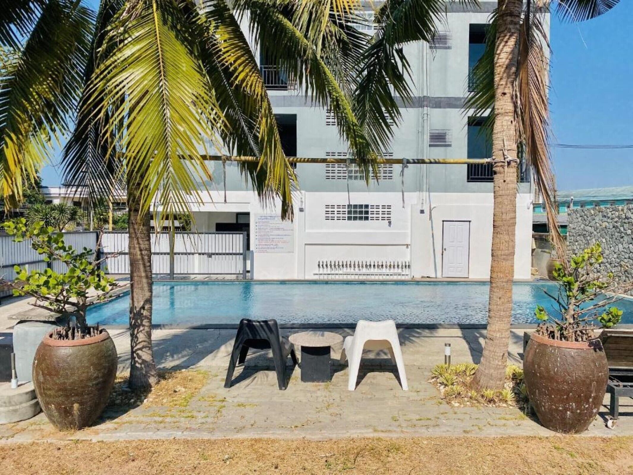 A pool area with two white plastic chairs and a table, surrounded by large potted plants and palm trees.