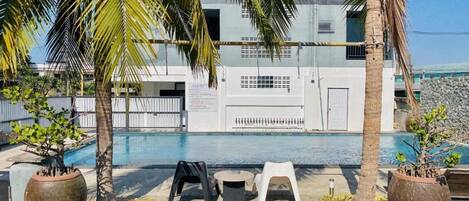 A pool area with two white plastic chairs and a table, surrounded by large potted plants and palm trees.